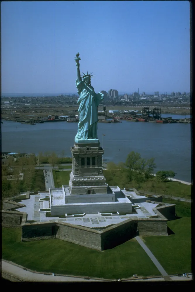 Aerial view of a tall green statue holding a torch aloft, standing on a star-shaped base on an island